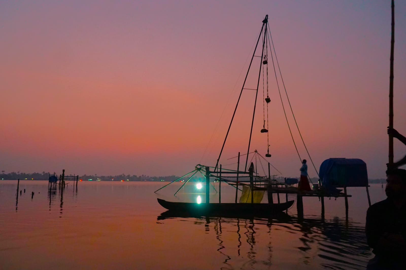 A fishing boat and the frame of a net silhouetted on still water under an orange sunset.