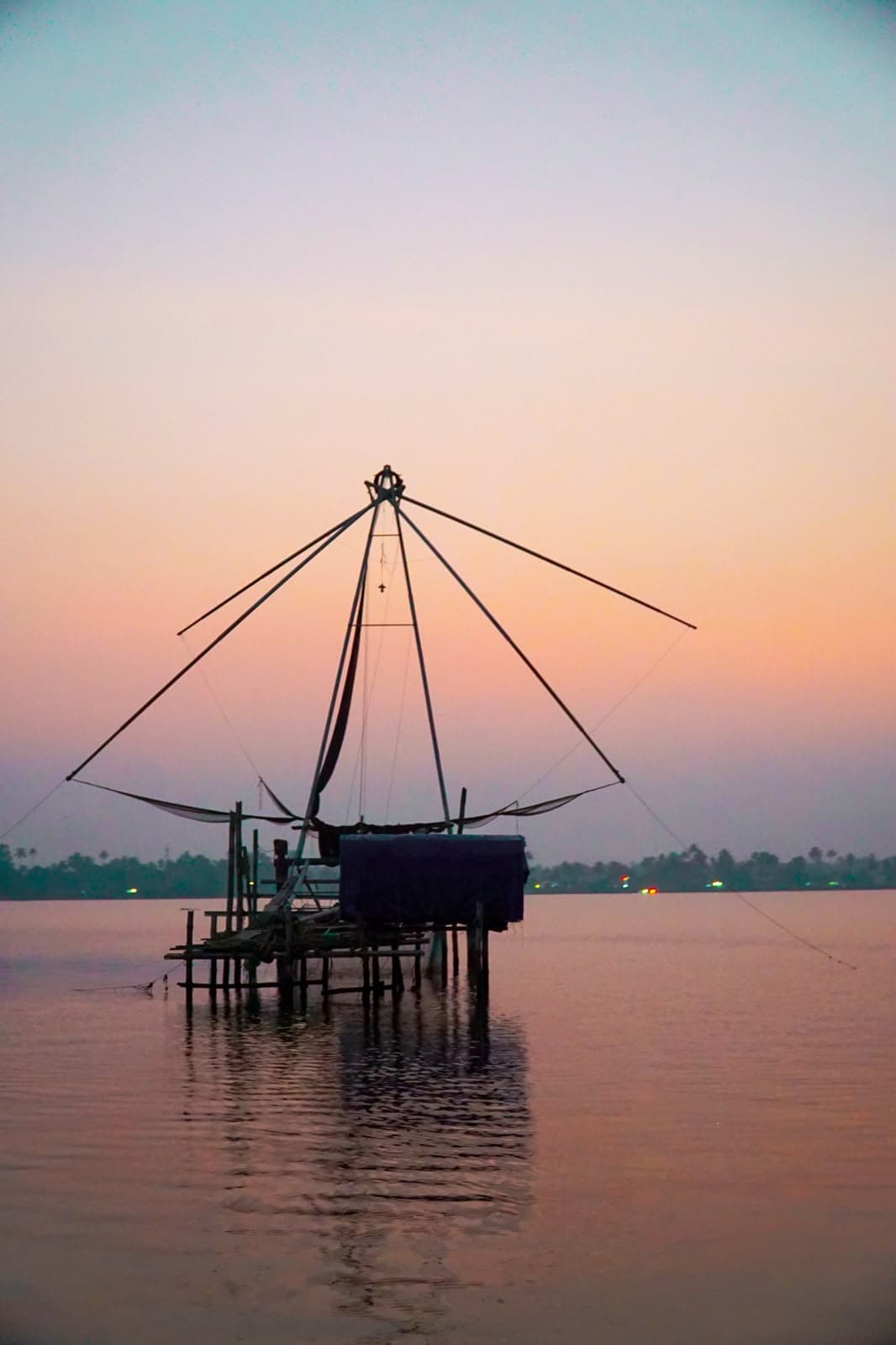 A Chinese fishing net standing on stilts in calm water beneath a pale pastel sky.