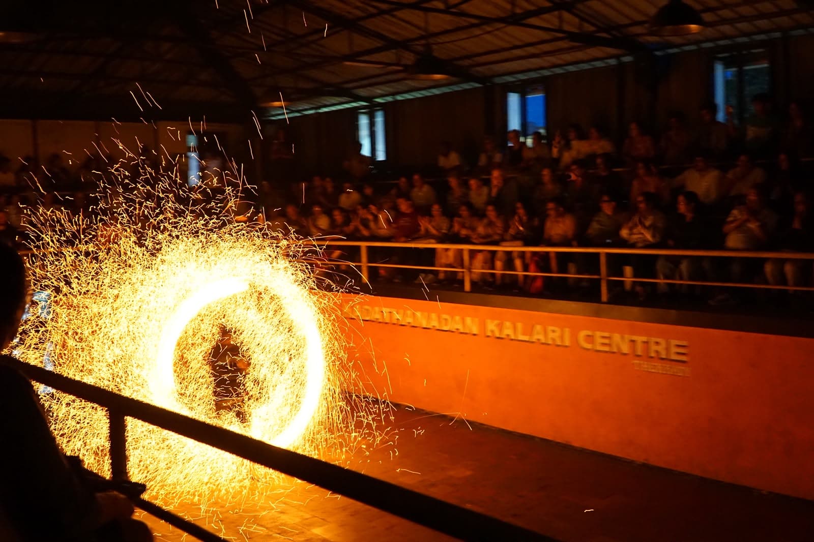 A Kalaripayattu performer spinning a blazing ring of fire before a seated audience.