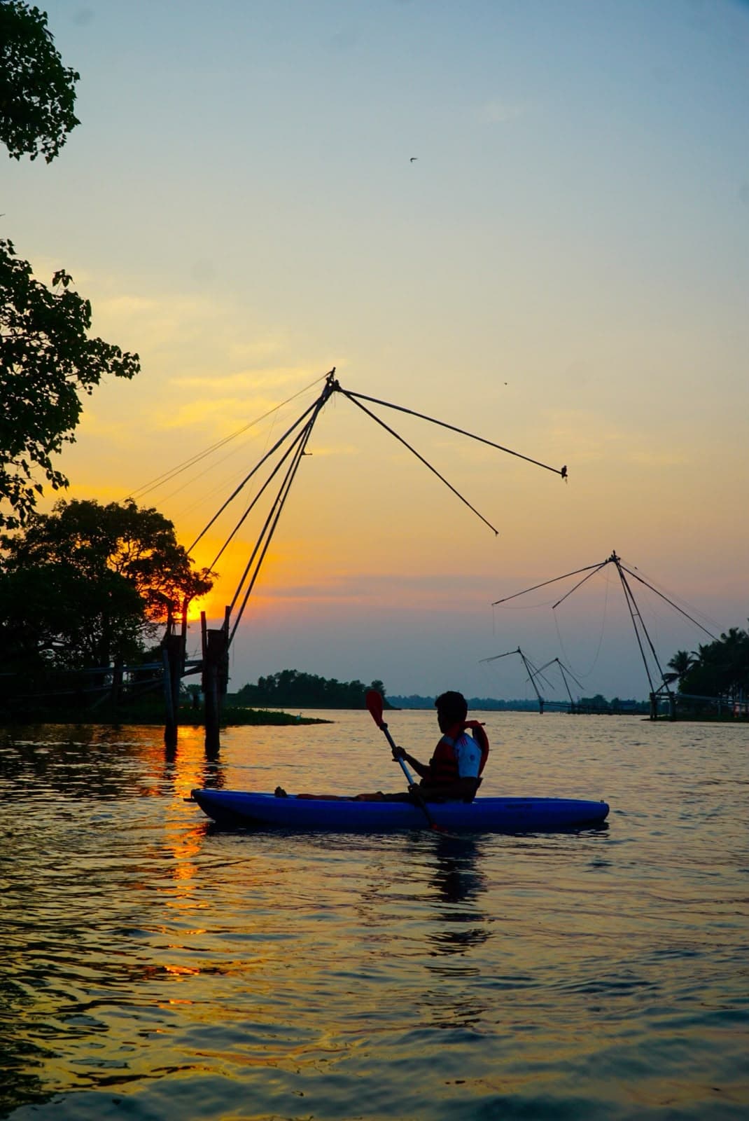 Two people in a blue kayak crossing calm water past fishing nets at sunset.