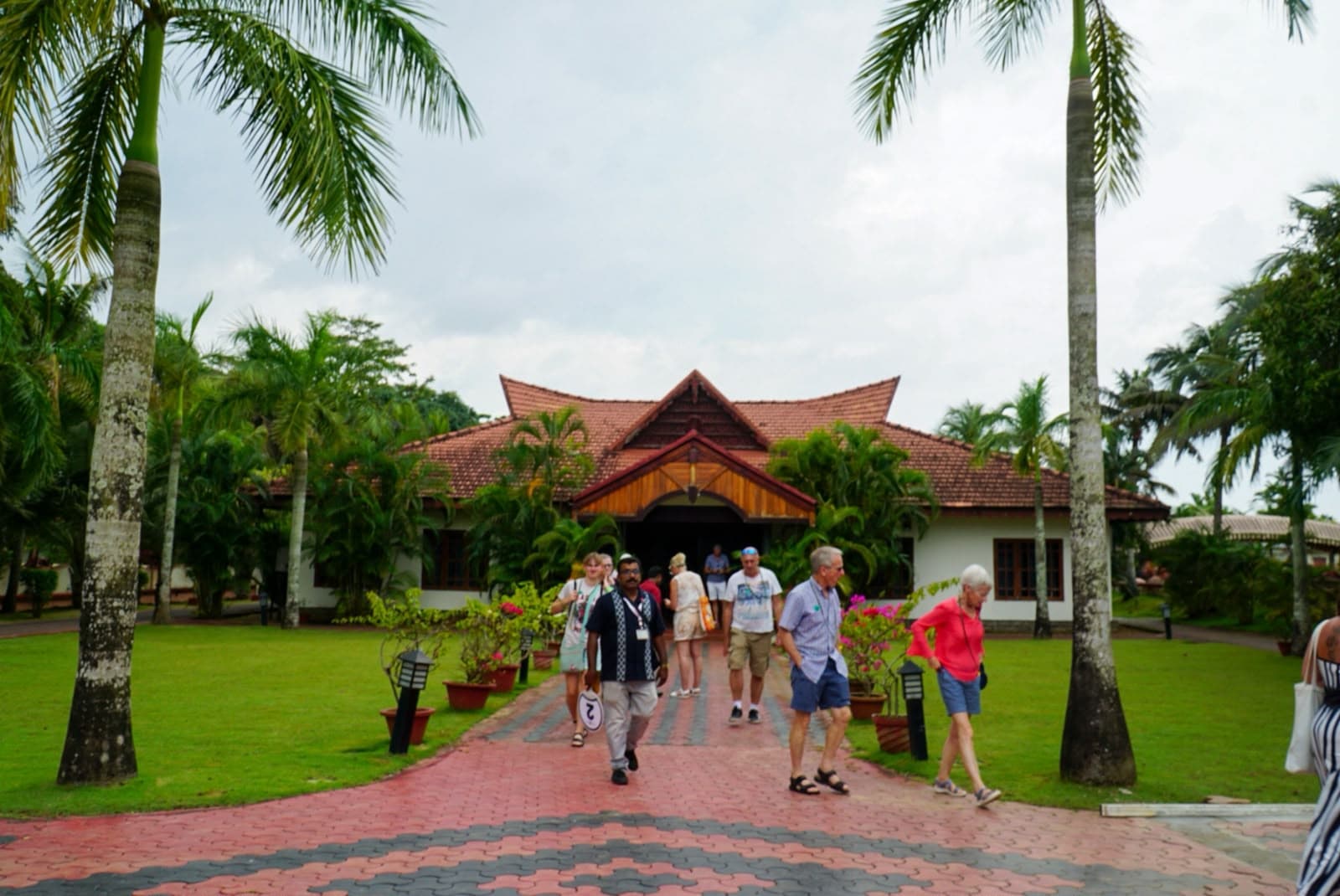 Travellers walking a brick path towards a tile-roofed Kerala building between tall coconut palms.