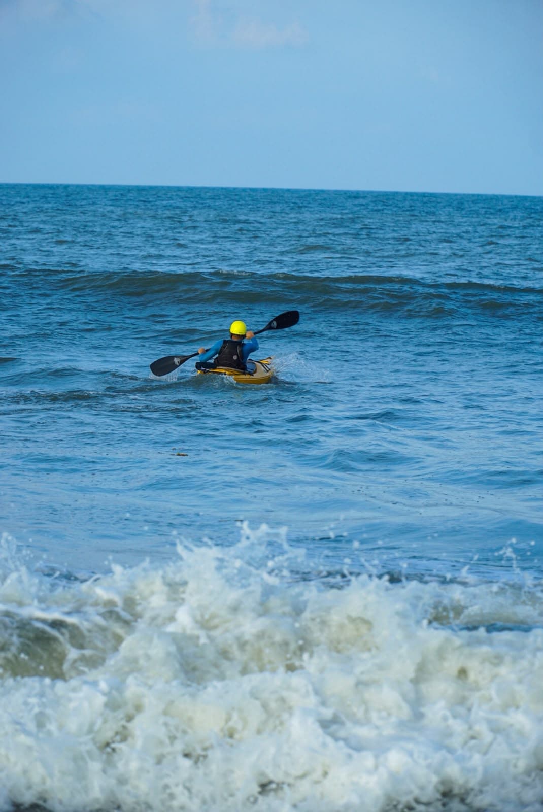 A kayaker paddling away from the shore across deep blue water and white foam.