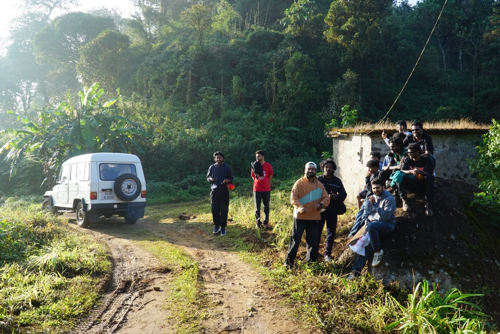 A jeep on a plantation track with a group resting beside a hut in the Idukki hills.